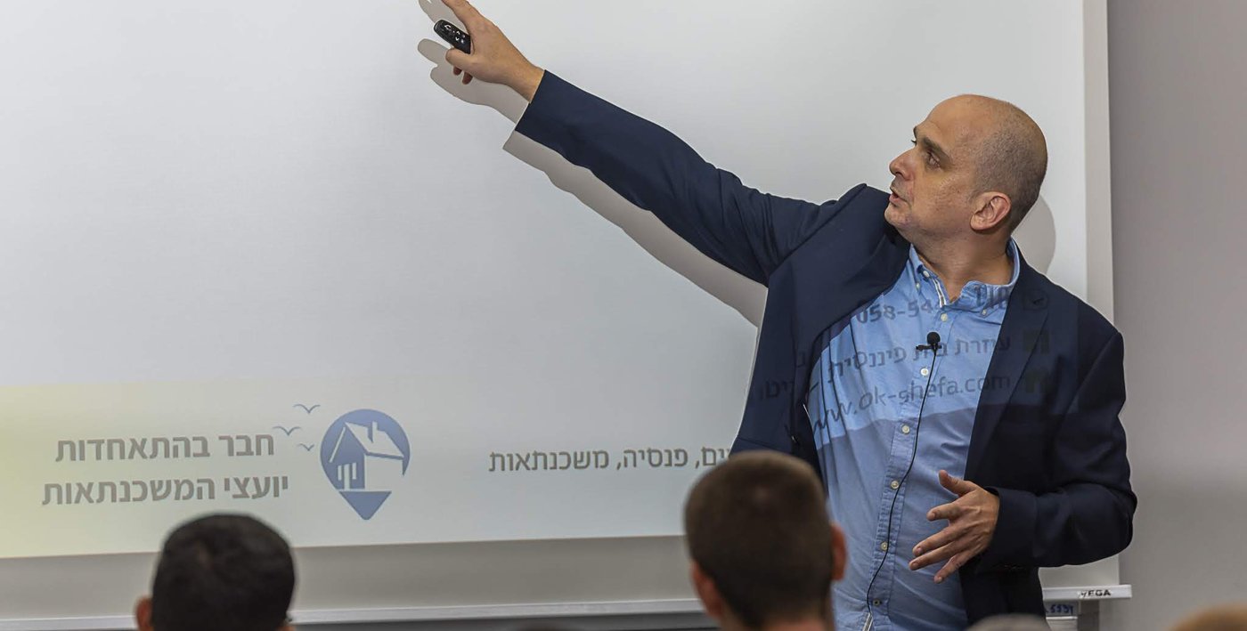 אורן מרצה מול קהל, ועל המסך הלוגו של התאחדות יועצי המשכנתאות